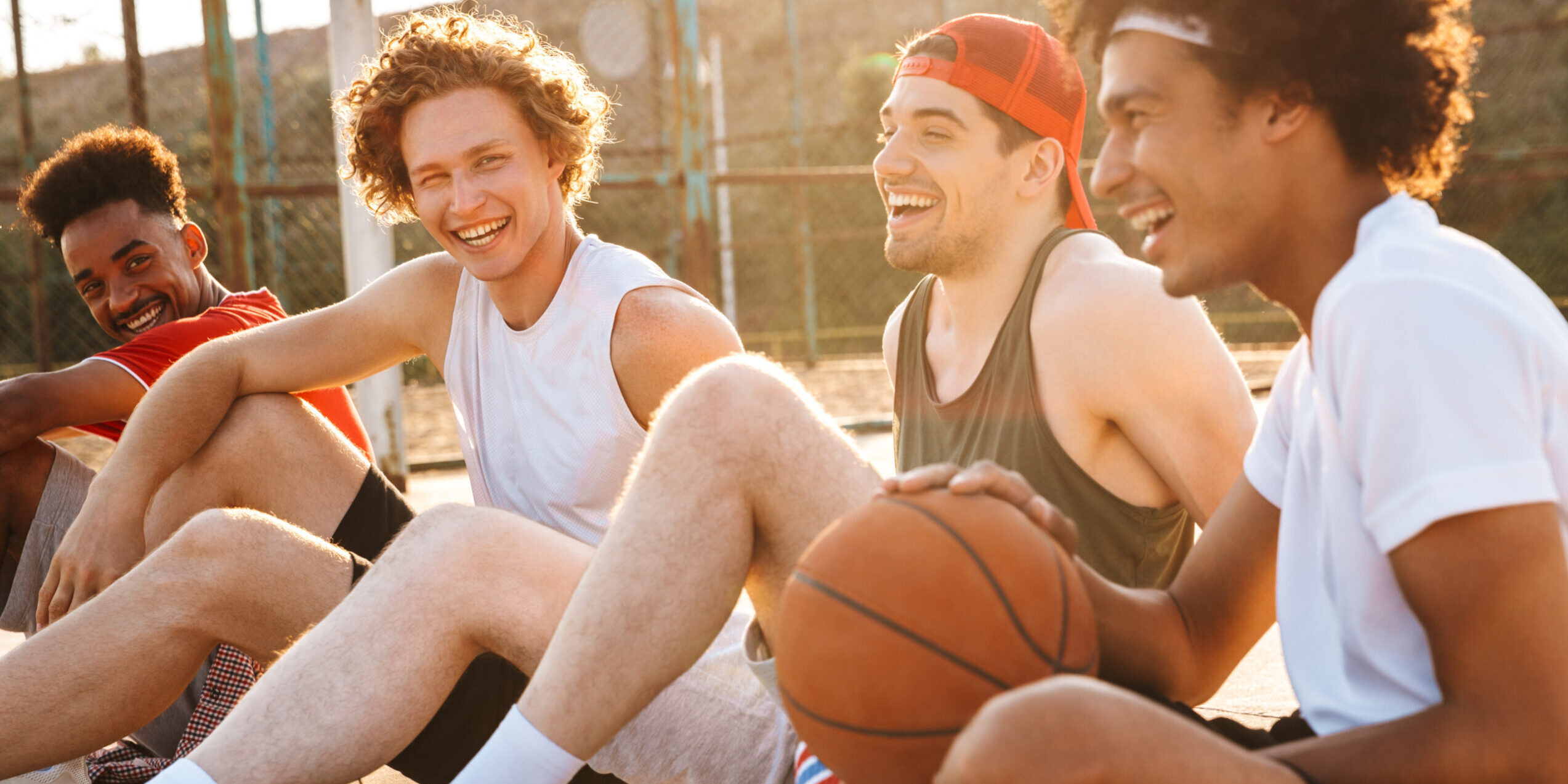 Young happy basketball players sitting at playground outdoor and watching game during summer sunny day
