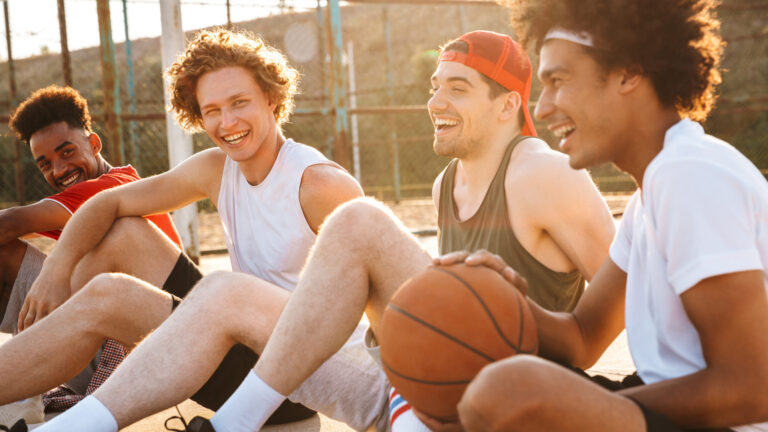 Young happy basketball players sitting at playground outdoor and watching game during summer sunny day